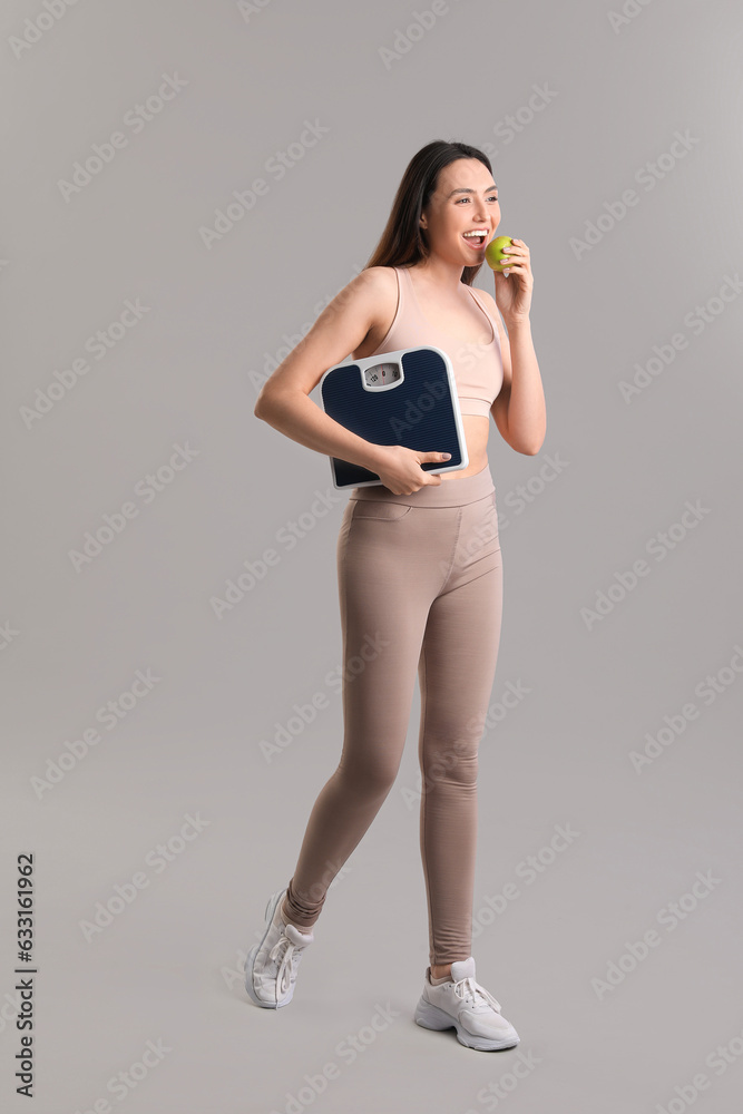 Sporty young woman with scales eating apple on grey background