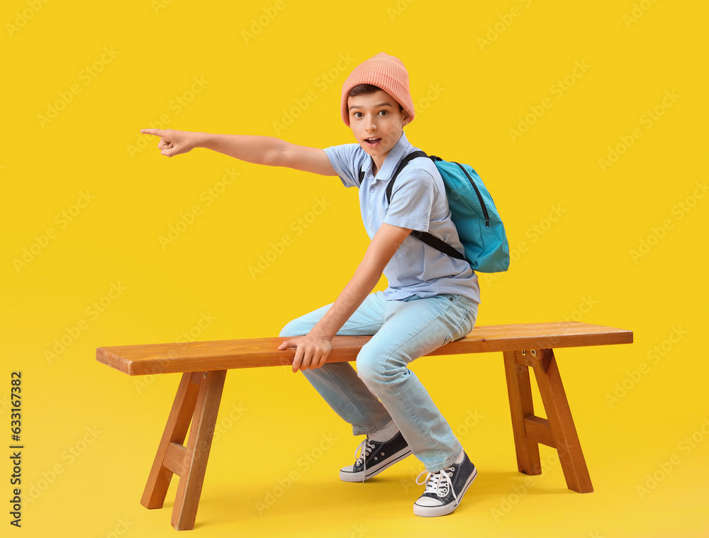 Portrait of shocked schoolboy sitting on bench and pointing somewhere on orange background