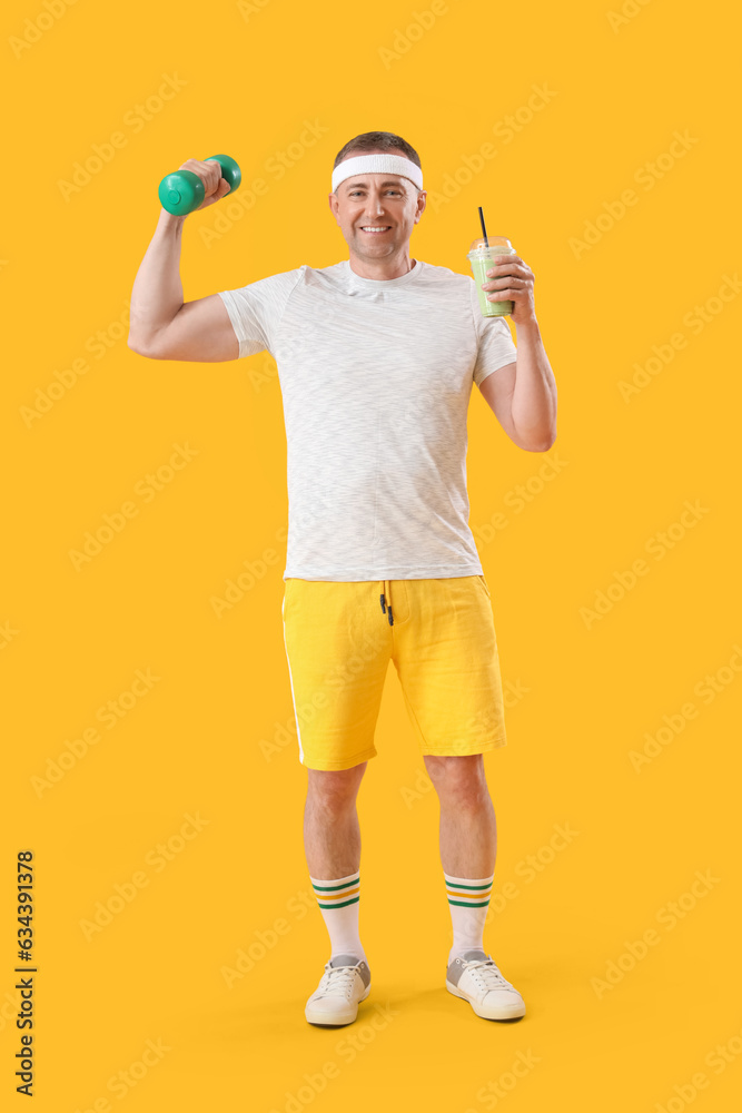 Sporty mature man with glass of vegetable smoothie and dumbbell on yellow background