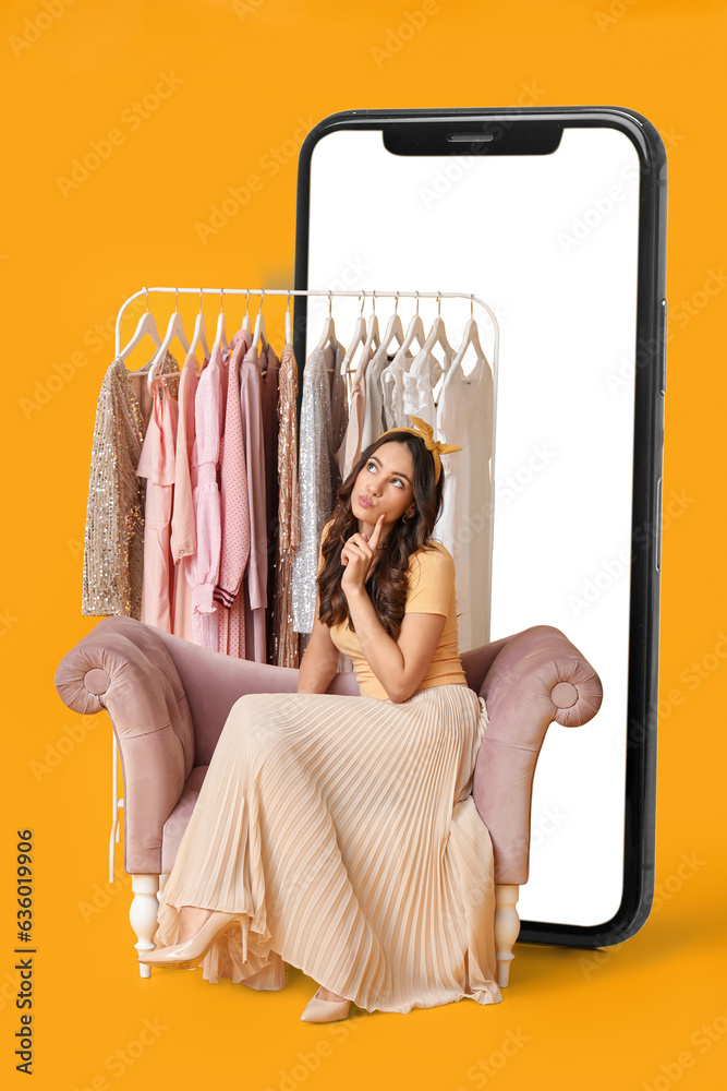 Thoughtful young woman sitting near rack with stylish clothes and big mobile phone on yellow backgro