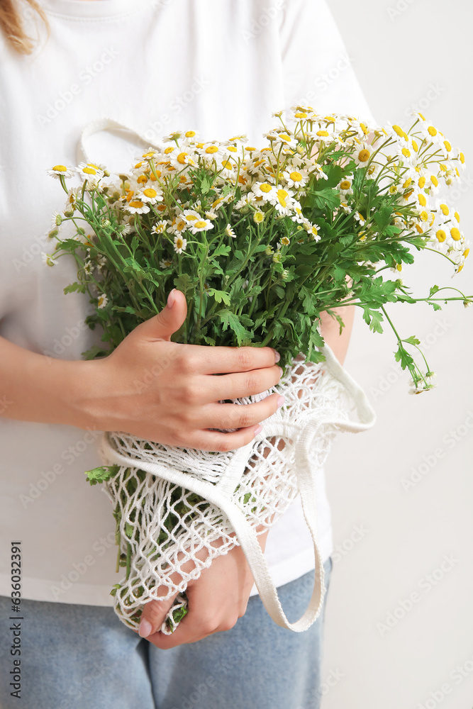 Woman holding mesh bag with beautiful chamomile flowers on light background, closeup