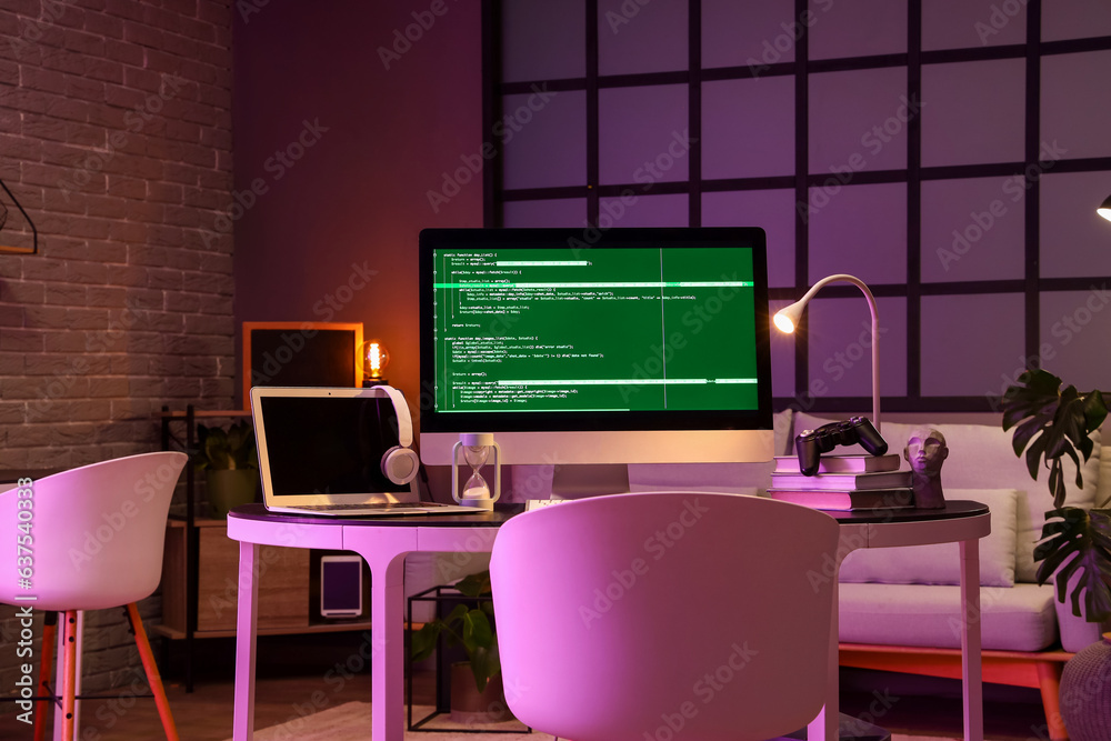 Programmers workplace with computer, laptop and glowing lamp in dark office