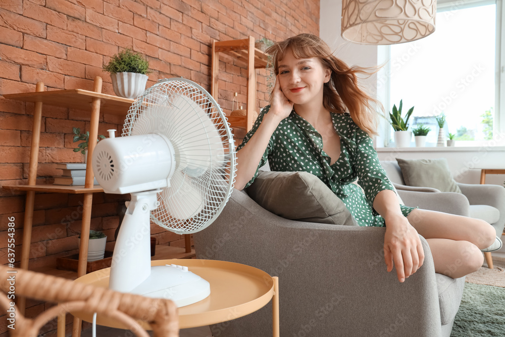 Young woman with blowing electric fan sitting on sofa at home