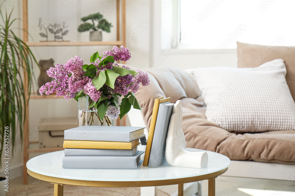 Stylish holder for books with vase of beautiful lilac flowers on coffee table in living room