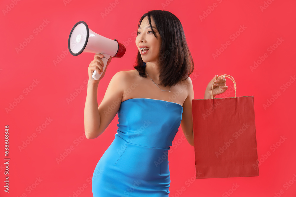 Beautiful happy stylish Asian woman with shopping bag and megaphone on red background