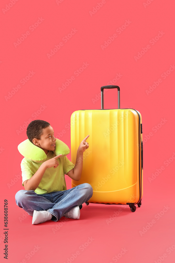 Little African-American boy sitting on floor and pointing at suitcase on red background