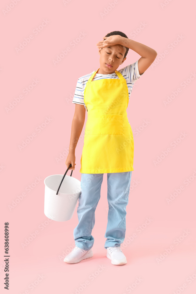 Tired African-American boy in apron with bucket on pink background