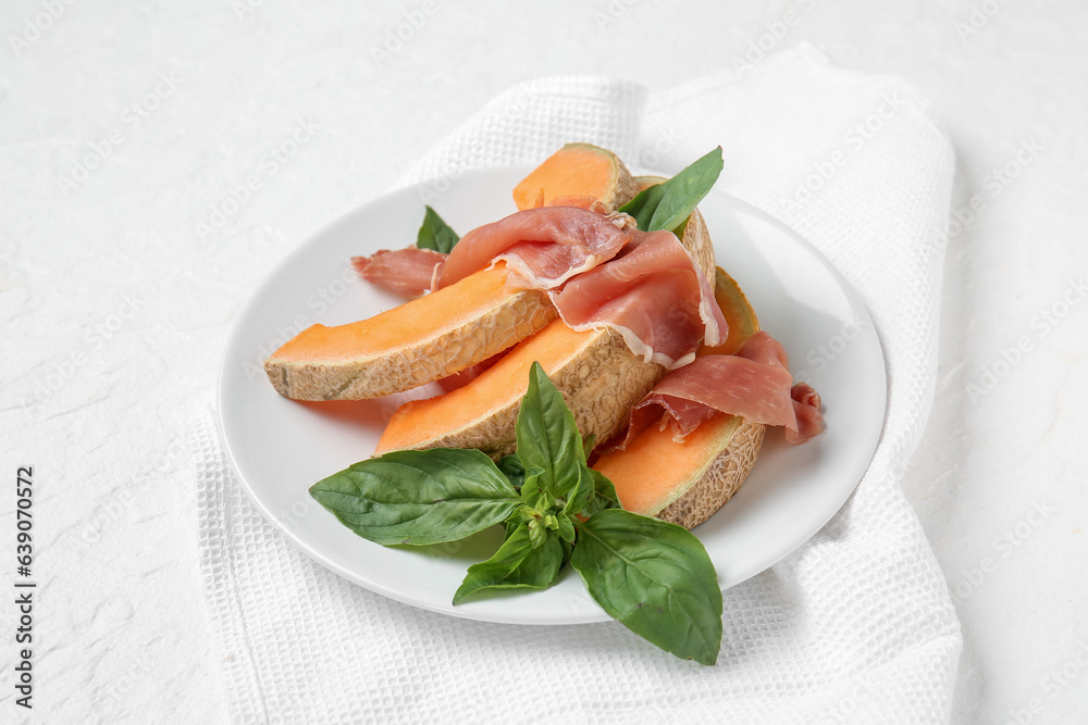 Plate with tasty melon, prosciutto and basil on light background