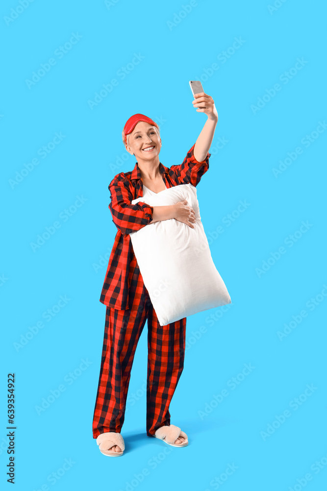 Mature woman in pajamas with pillow taking selfie on blue background