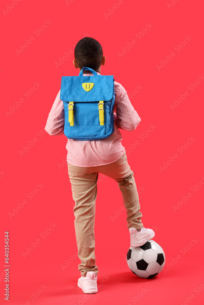 Little African-American schoolboy with soccer ball and backpack on red background, back view