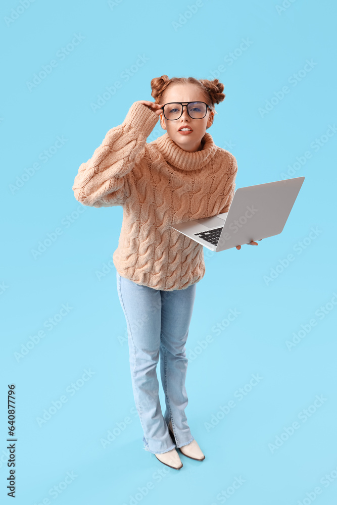 Thoughtful beautiful young woman in warm sweater and eyeglasses using laptop on blue background