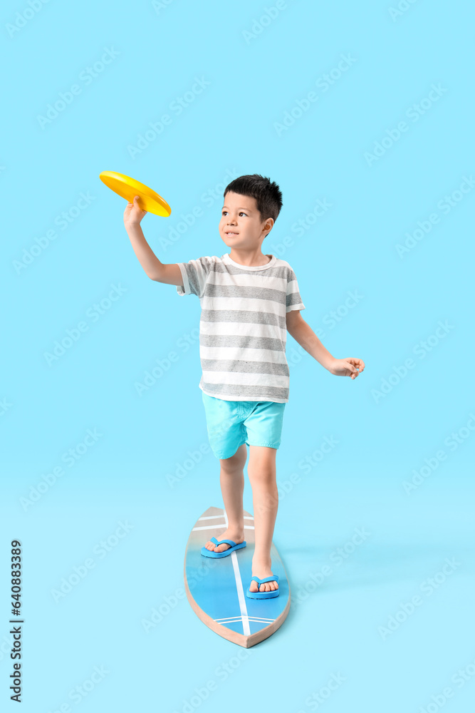 Cute little Asian boy standing on surfboard and playing with flying disc against blue background