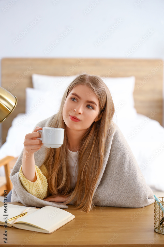 Thoughtful young woman with cup of coffee at table in bedroom