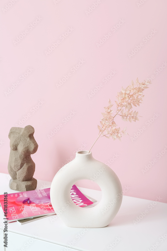 Vase with dried twig, figurine and magazines on dresser near pink wall