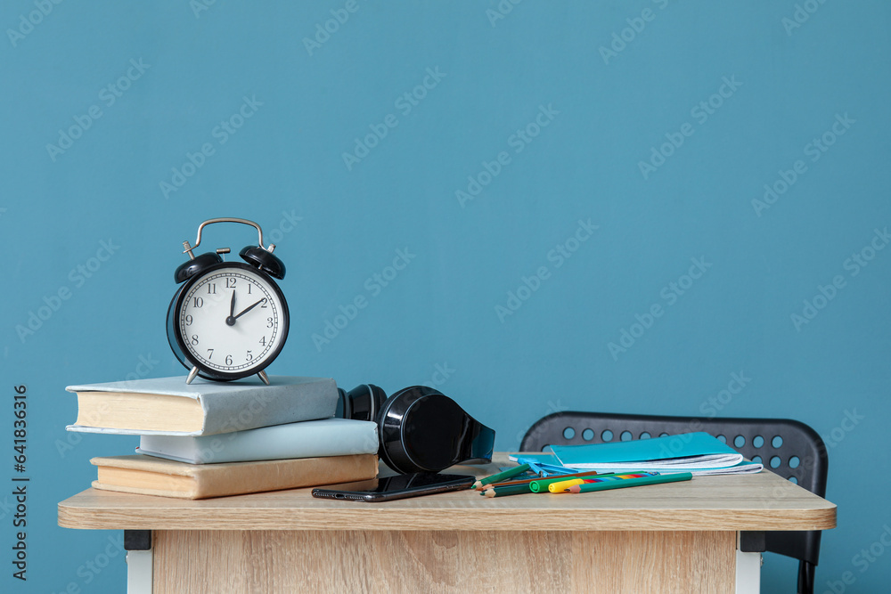 Modern school desk with alarm clock, headphones and stationery in room near blue wall