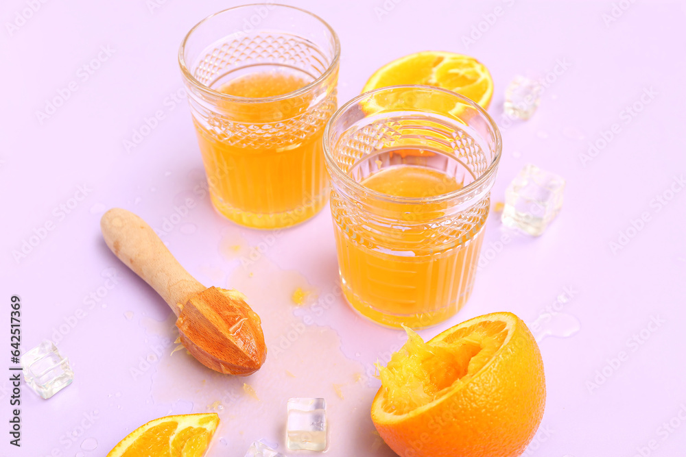 Glasses of orange juice with ice cubes and juicer on lilac background