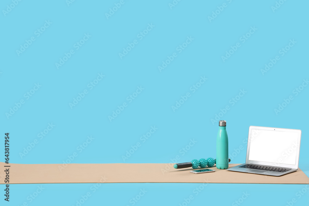 Fitness mat with laptop, mobile phone, bottle and massage roller on blue background