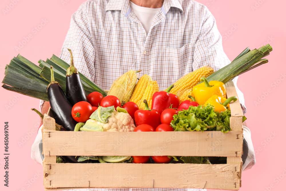 Mature male farmer with wooden box full of different ripe vegetables on pink background, closeup