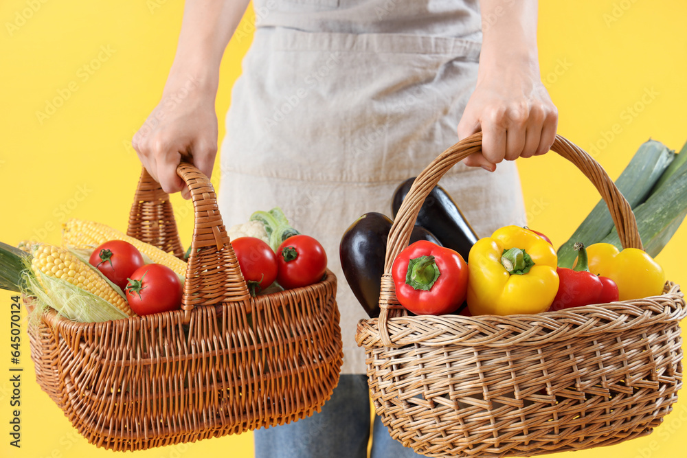 Young female farmer with wicker baskets full of different ripe vegetables on yellow background, clos