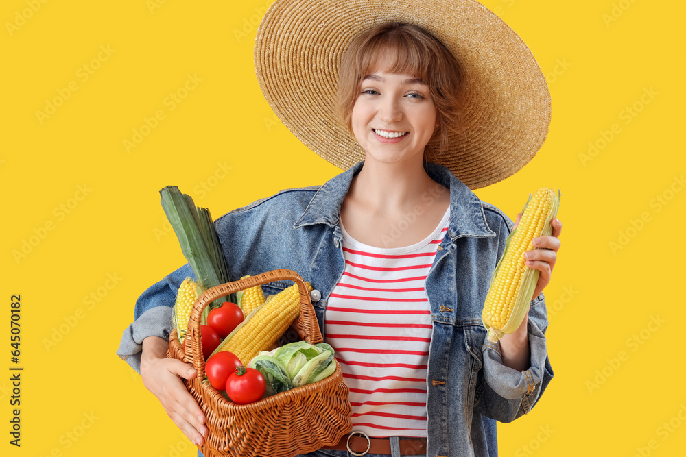 Young female farmer with corn and wicker basket full of different ripe vegetables on yellow backgrou