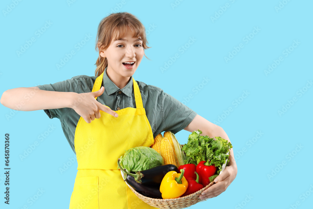 Happy young female farmer pointing at wicker basket full of different ripe vegetables on blue backgr