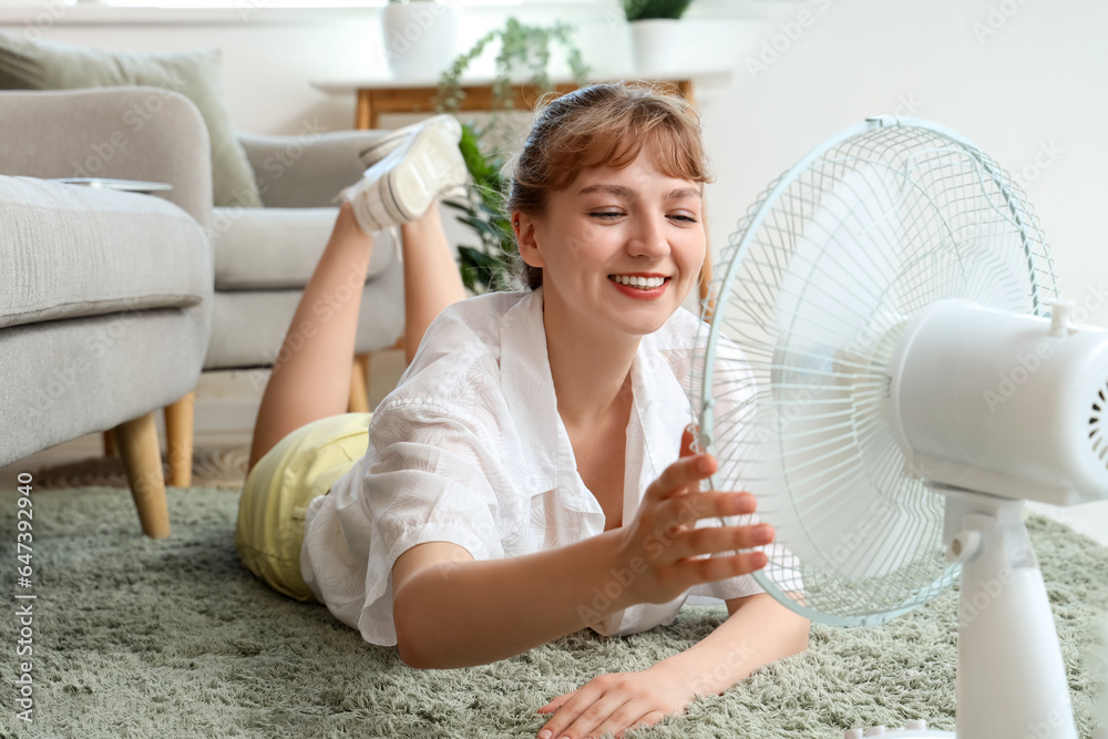 Young woman with blowing electric fan lying on floor at home