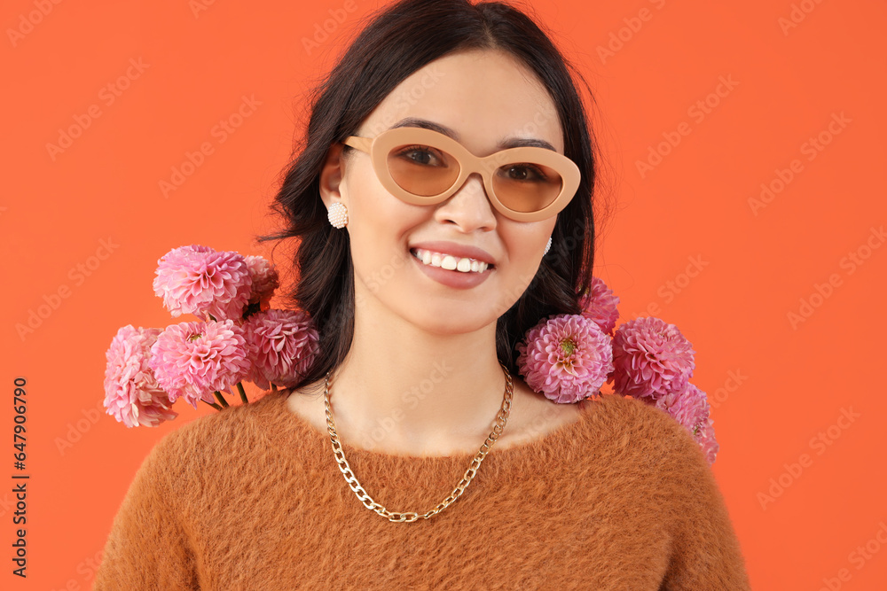 Stylish young Asian woman in sunglasses with flowers on orange background, closeup