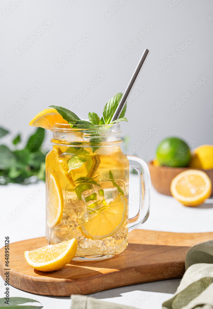 Lemonade drink or cold tea with fresh lemon, mint and ice in a jar. Refreshing citrus mojito cocktail on a wooden board on a  light background with fresh fruits and shadows.