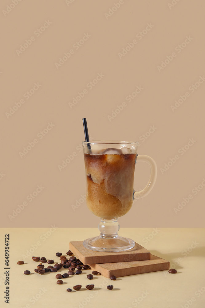 Glass of ice coffee with straw and beans on yellow table near beige wall