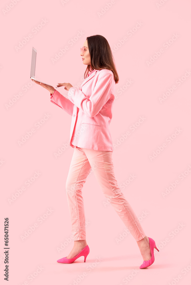 Young woman in stylish suit using laptop on pink background