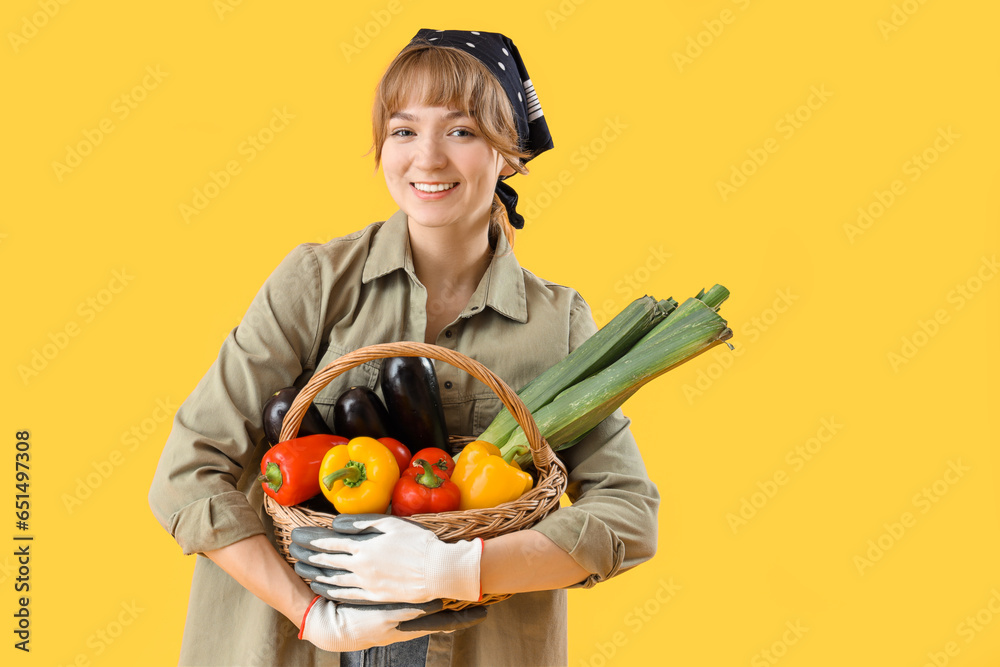 Happy young female farmer with wicker basket full of different ripe vegetables on yellow background