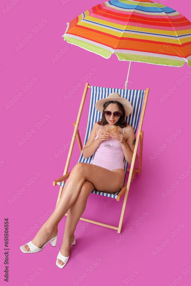 Beautiful young woman with umbrella and glass of tasty cocktail sitting on deckchair against purple background