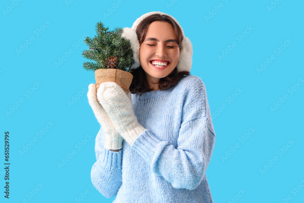 Young woman in warm clothes with small Christmas tree on blue background