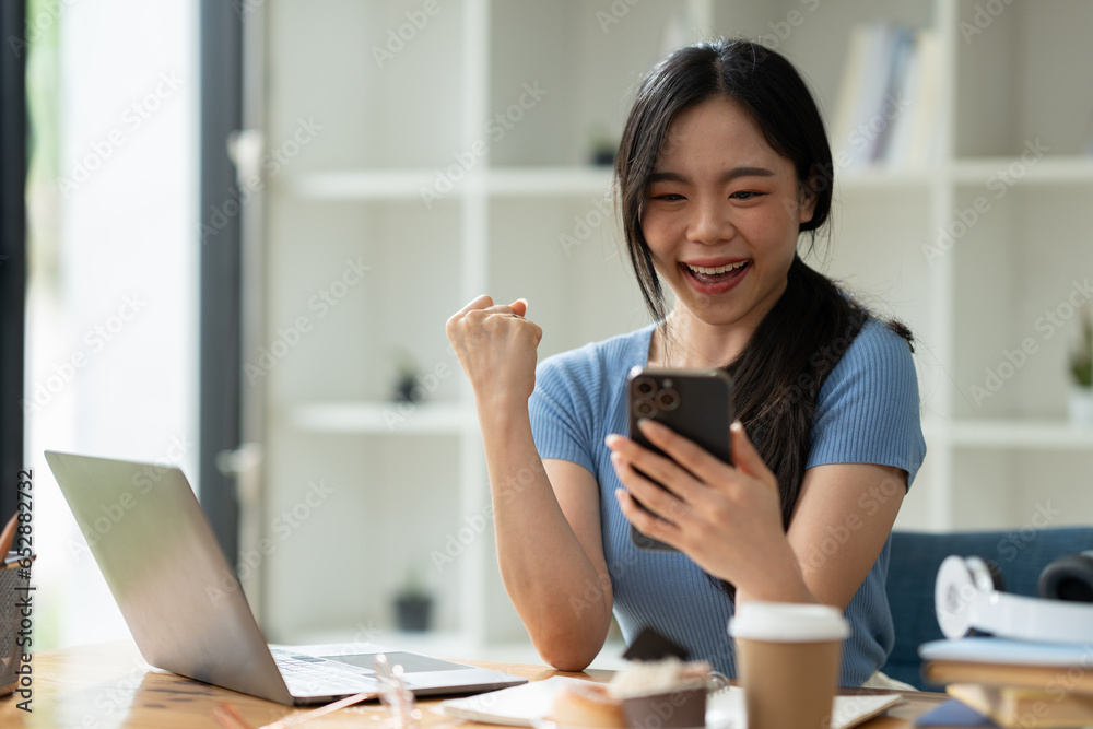 Asian woman sitting at work happily, smiling, delighted with business success feedback data from smartphone and data on laptop. while sitting on a desk in his office at home