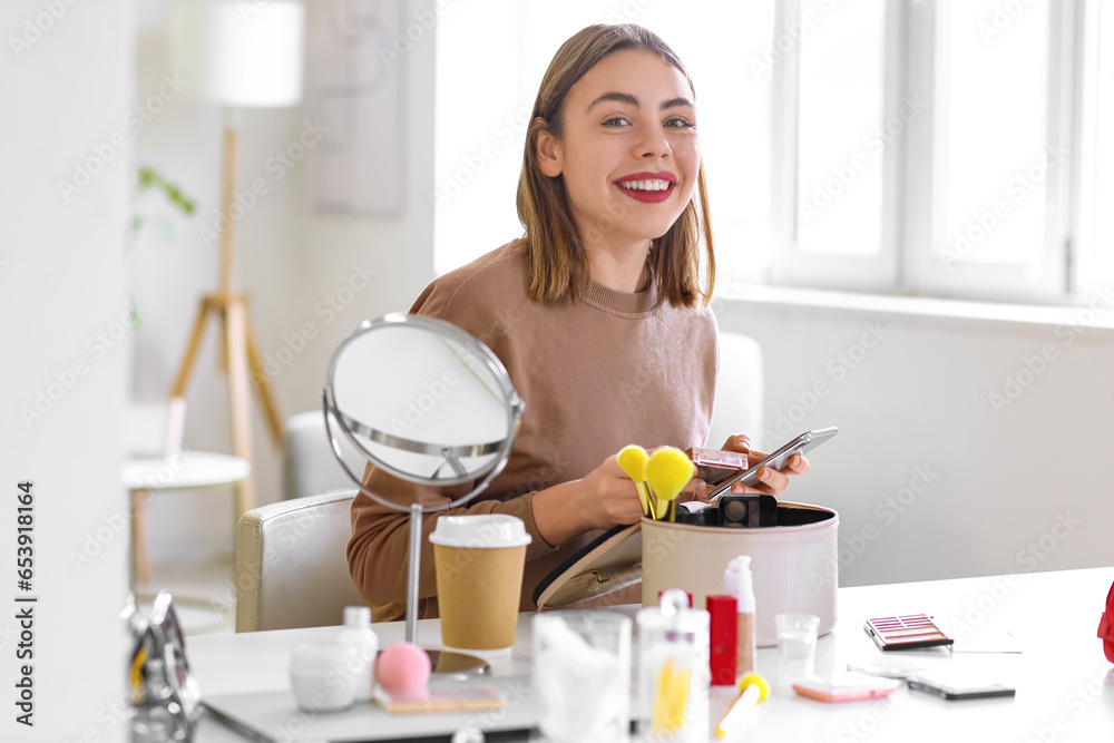 Young woman with makeup product and mobile phone at home