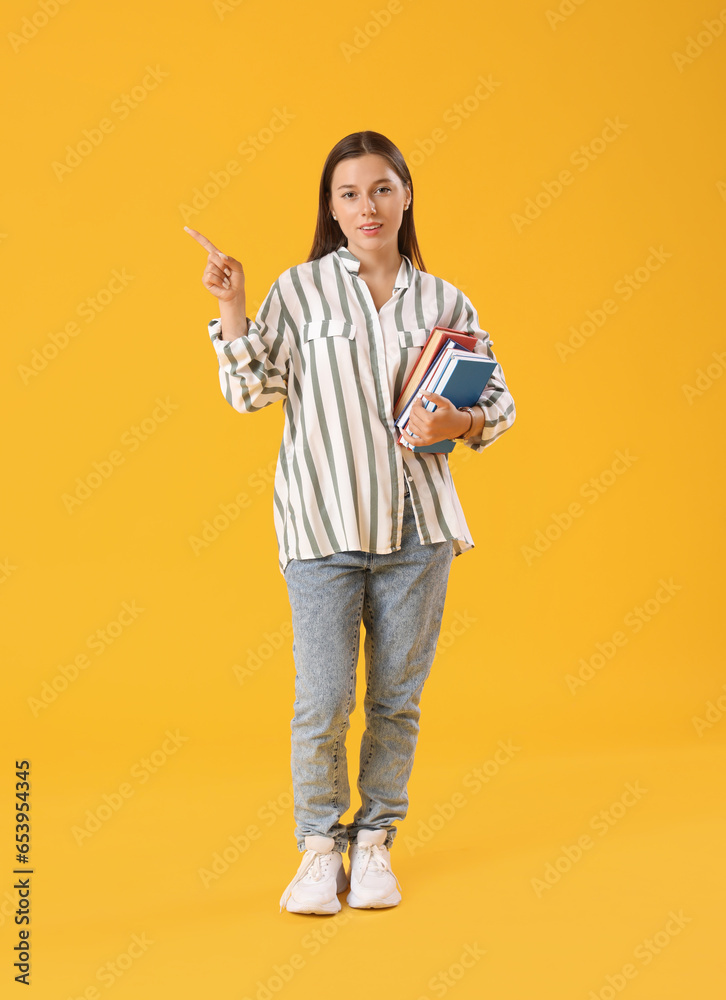 Happy female student with many books pointing at something on yellow background