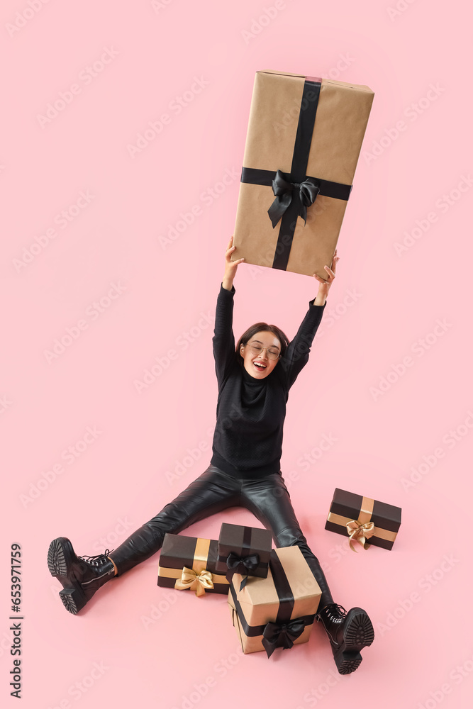 Young Asian woman with gifts sitting on pink background. Black Friday sale