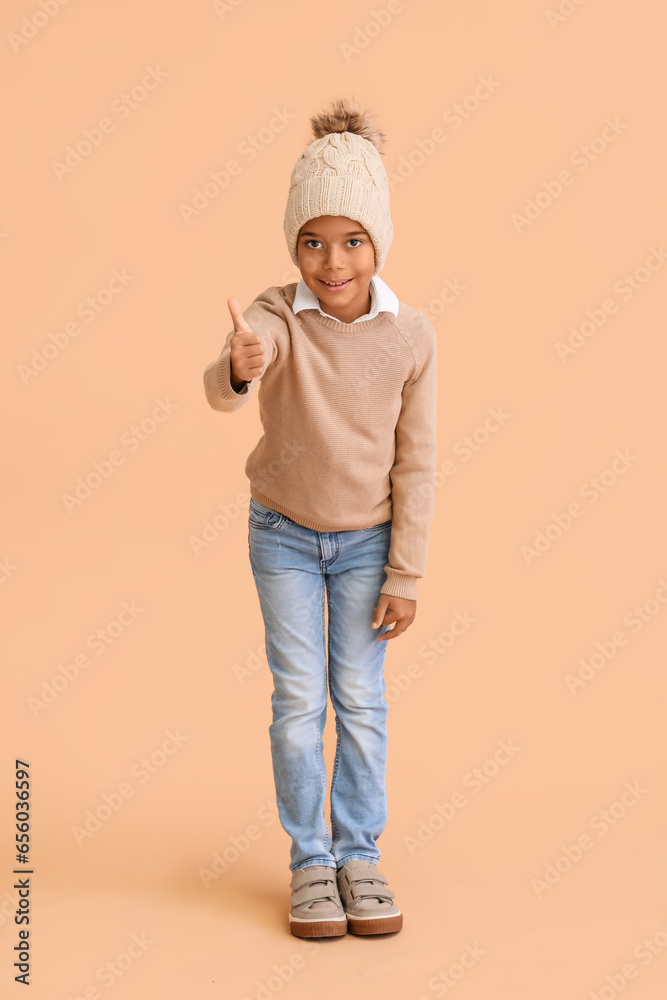 Cute African-American boy in warm winter clothes showing thumb-up on beige background