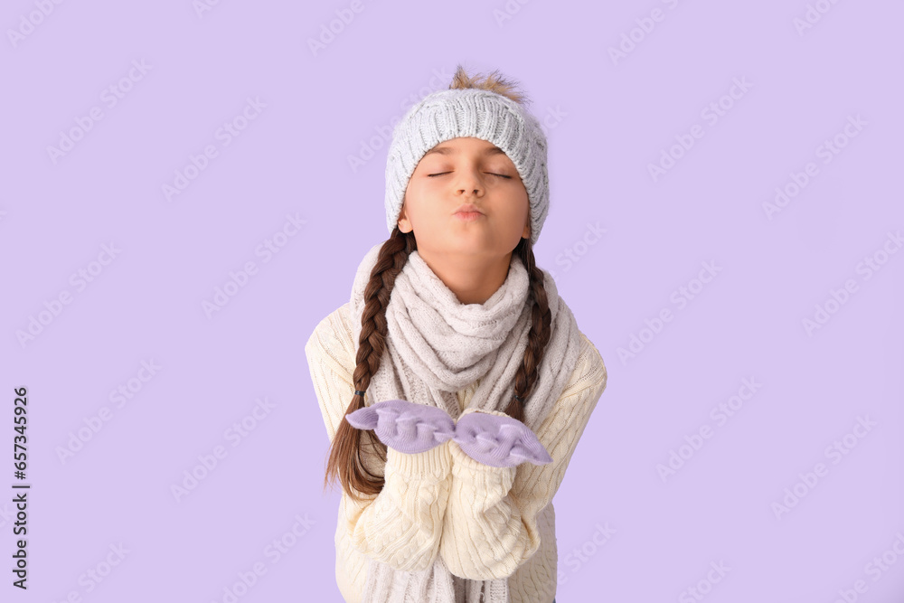 Cute little girl in winter clothes blowing kiss on lilac background