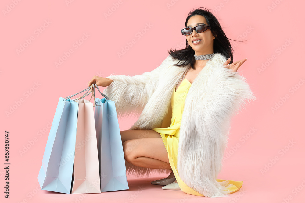 Happy beautiful young Asian woman in sunglasses with shopping bags sitting on pink background