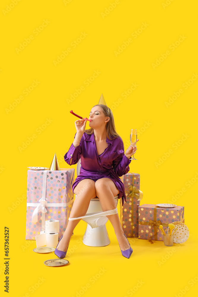 Young woman with champagne and party blower sitting on toilet bowl against yellow background