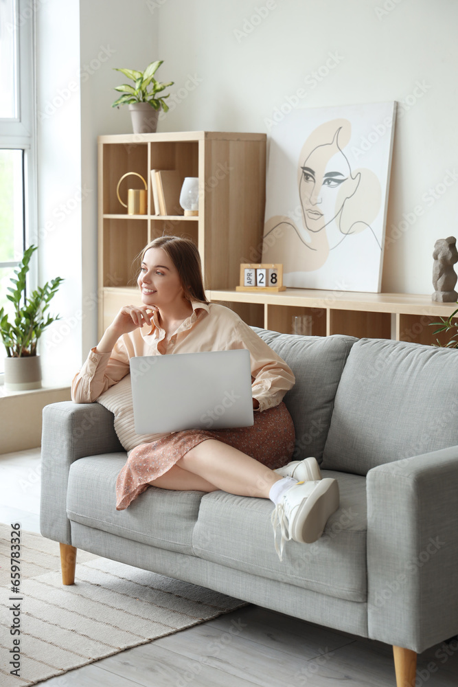 Pretty young woman sitting on grey sofa and using modern laptop in light living room