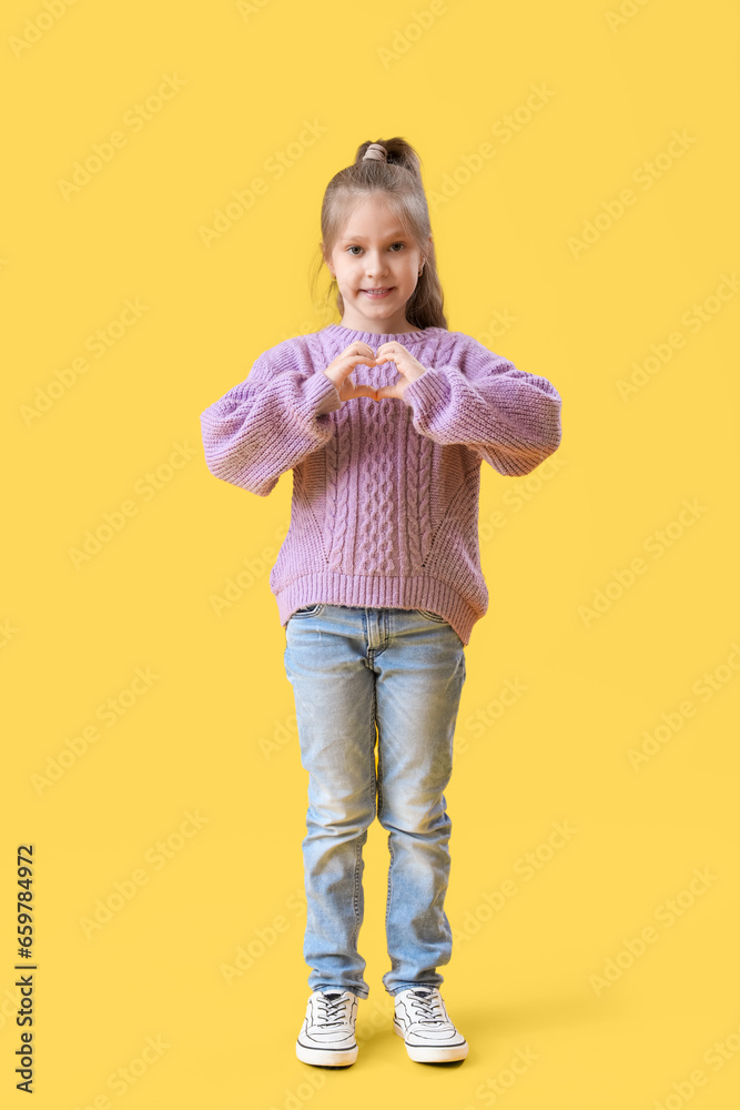 Cute little girl in knitted sweater making heart with her hands on yellow background