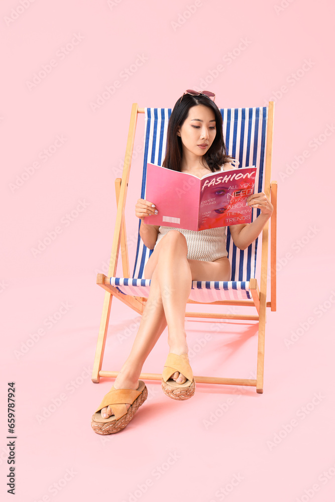 Beautiful Asian woman reading magazine in deck chair on pink background