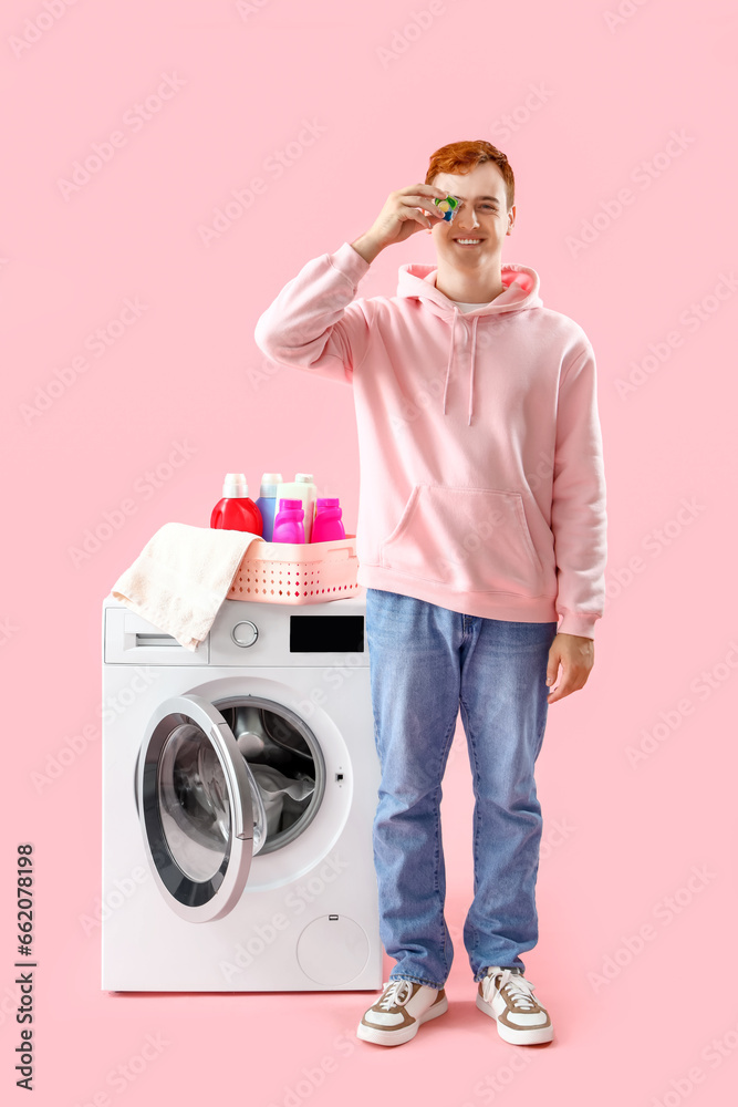 Young man with laundry capsule near washing machine on pink background