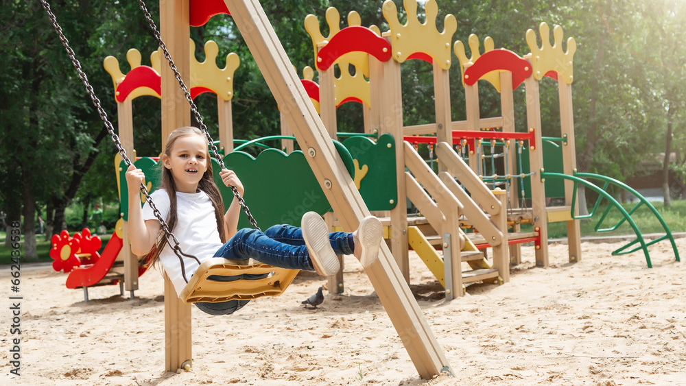 Little girl rides on a swing in an outdoor playground. Children play in the courtyard of a school or kindergarten. Healthy summer fun for kids.