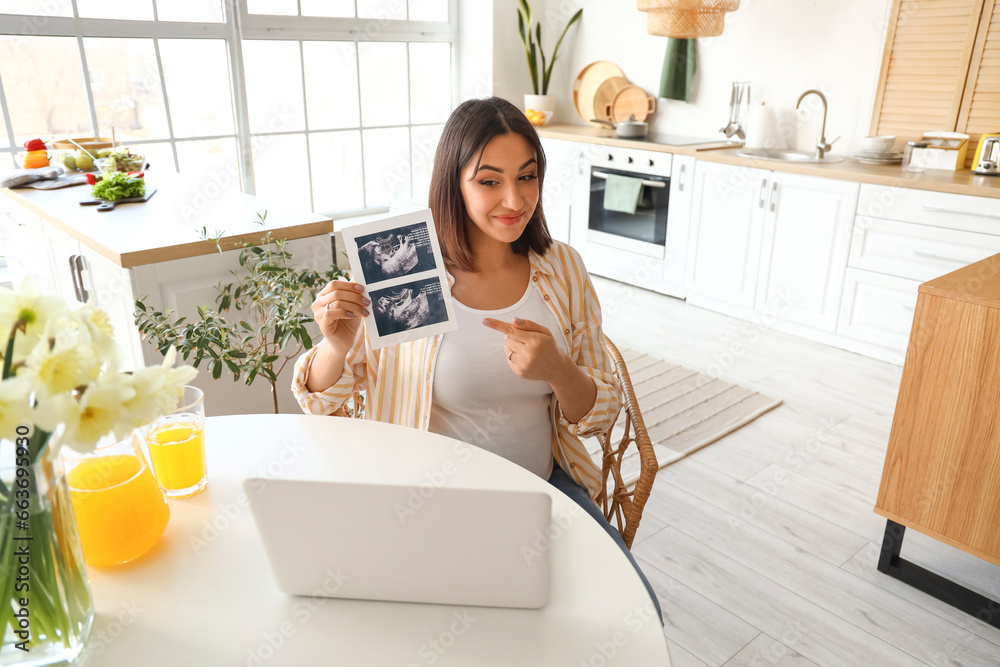 Young pregnant woman with sonogram image video chatting at table in kitchen