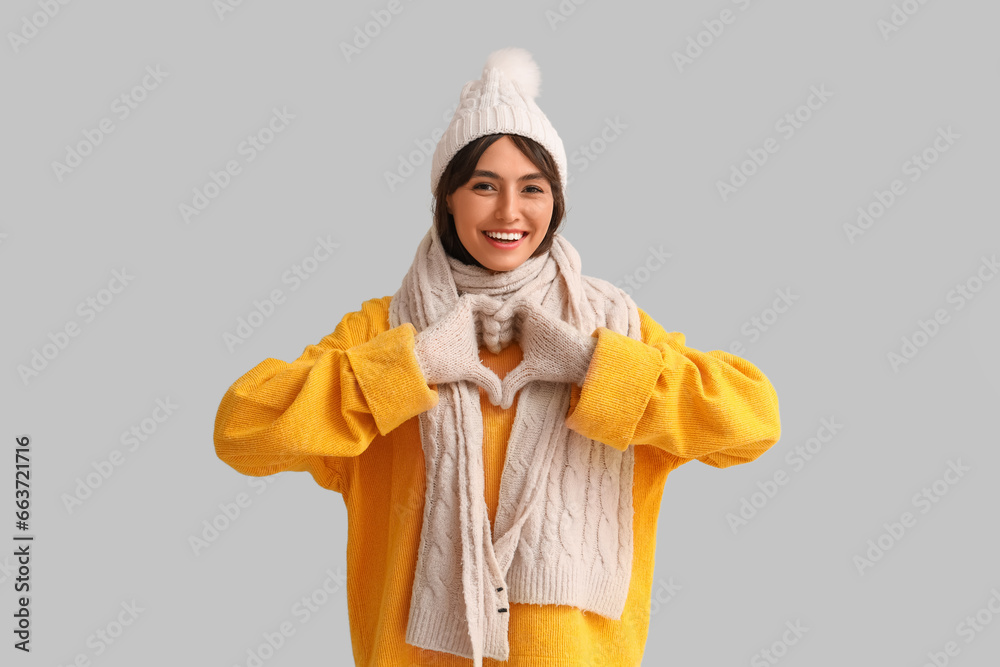Young woman in warm clothes making heart gesture on light background