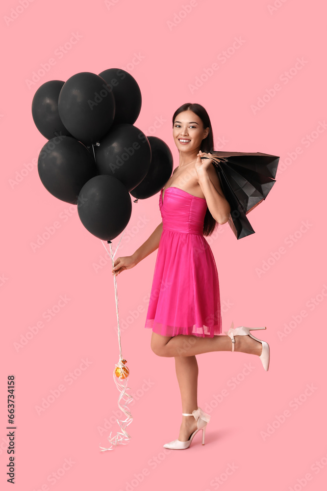 Beautiful young Asian woman with shopping bags and balloons on pink background. Black Friday sale
