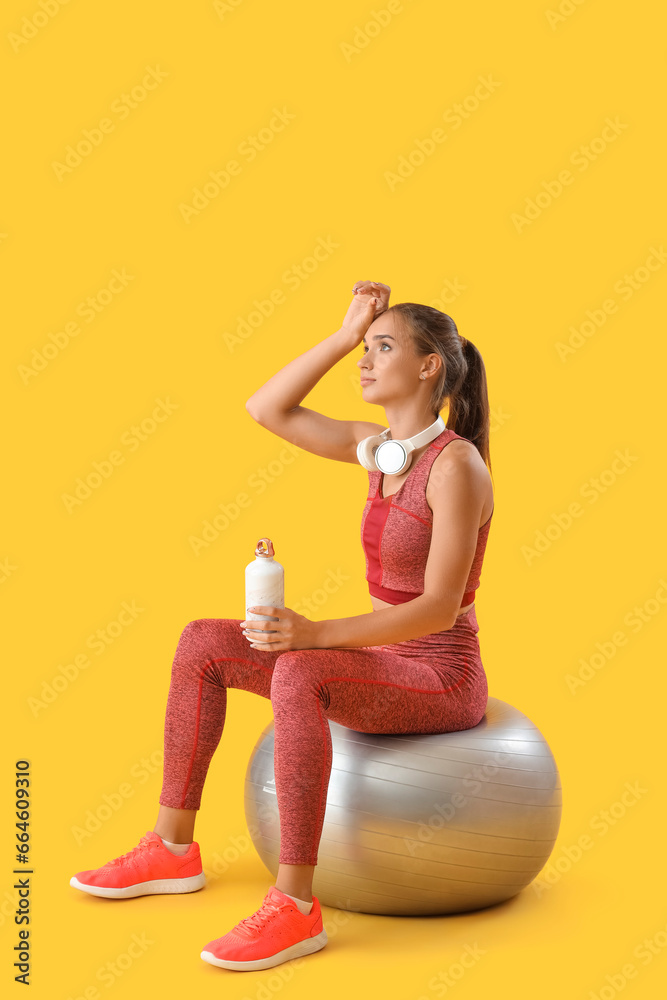 Sporty young woman with bottle of water and fitball resting after training on yellow background